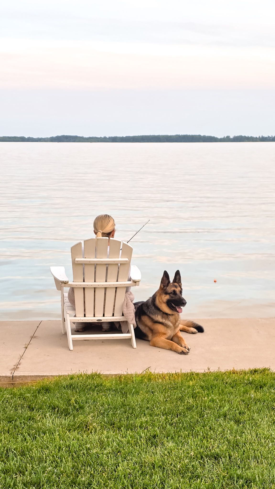 Person fishing from the Grand Lake shoreline beside a German shepherd