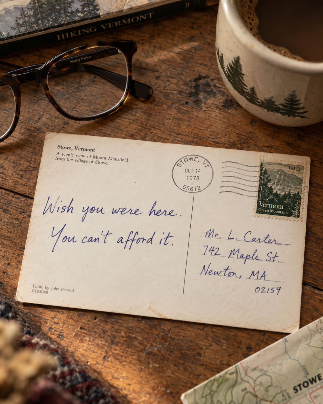 A vintage Vermont postcard on a wooden table beside reading glasses and coffee, handwritten in blue ink: &ldquo;Wish you were here. You can&rsquo;t afford it.&rdquo; Postmarked Stowe, Vermont, 1978.