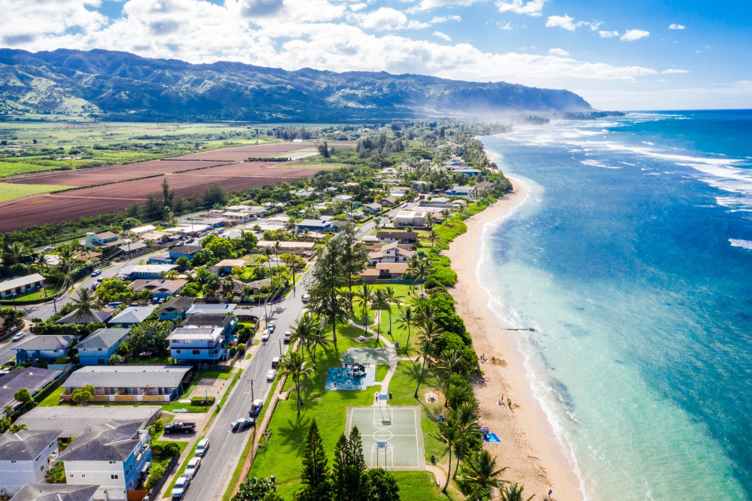 Aerial of a Waialua neighborhood, beach park and farm fields below a mountain ridge