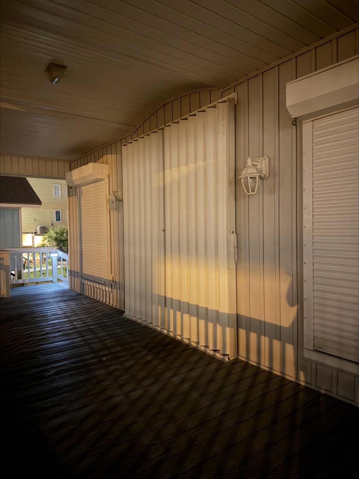 Accordion shutters and roll-down shutters closed along a covered wooden walkway in evening light.