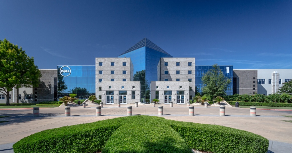 Exterior view of the Dell corporate headquarters campus on a clear, sunny day, featuring modern glass and stone multi-story office buildings, a landscaped driveway with trimmed hedges, and the Dell logo visible on the facade.