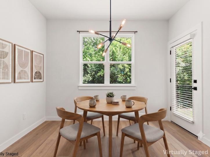 Virtually staged modern dining area featuring a round wood dining table with four upholstered chairs, contemporary Sputnik-style chandelier, light wood flooring, and large windows that fill the space with natural light and views of mature trees. A glass exterior door with blinds provides access to the backyard or patio, while neutral walls and minimalist artwork create a bright, inviting atmosphere ideal for everyday dining and entertaining.