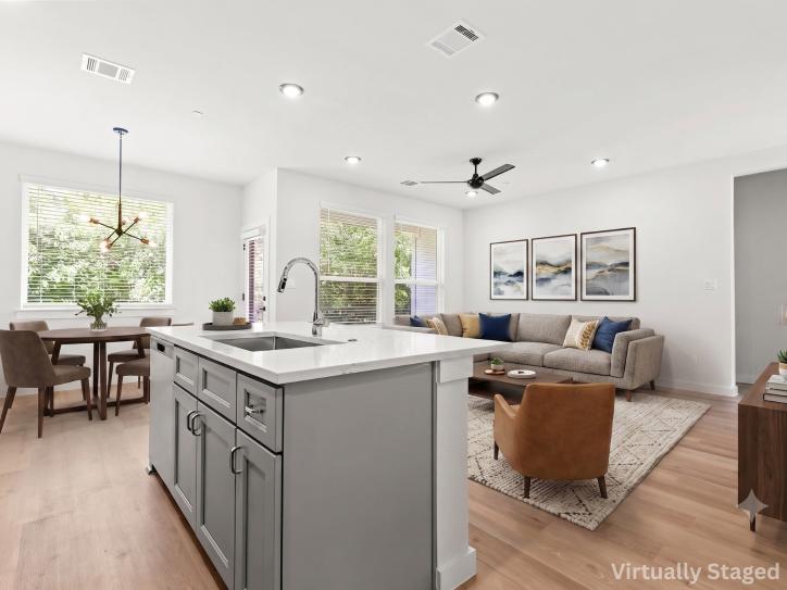 Virtually staged open-concept kitchen, dining, and living area featuring a spacious quartz island with an undermount sink, gray shaker-style cabinetry, stainless steel appliances, recessed lighting, and light wood flooring. The kitchen flows seamlessly into a bright dining area with large windows overlooking mature trees and a comfortable living room with contemporary furnishings, creating an ideal space for entertaining and everyday living.