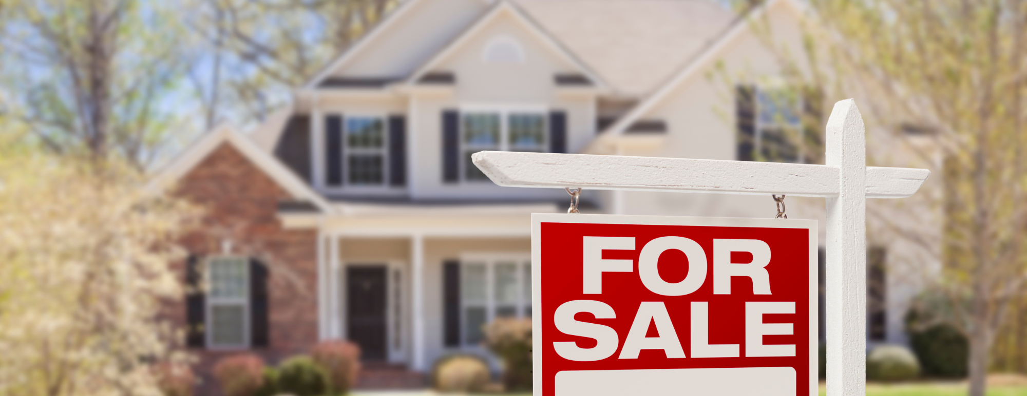 A red 'For Sale' sign with a white post stands prominently in front of a two-story home in Boise, Idaho, featuring mature landscaping. The house, softly out of focus in the background, highlights a welcoming atmosphere. The lush greenery and well-maintained landscaping create an inviting setting, perfect for potential buyers. This scene exemplifies the kind of homes Templeton Real Estate Group showcases in Boise's thriving market.