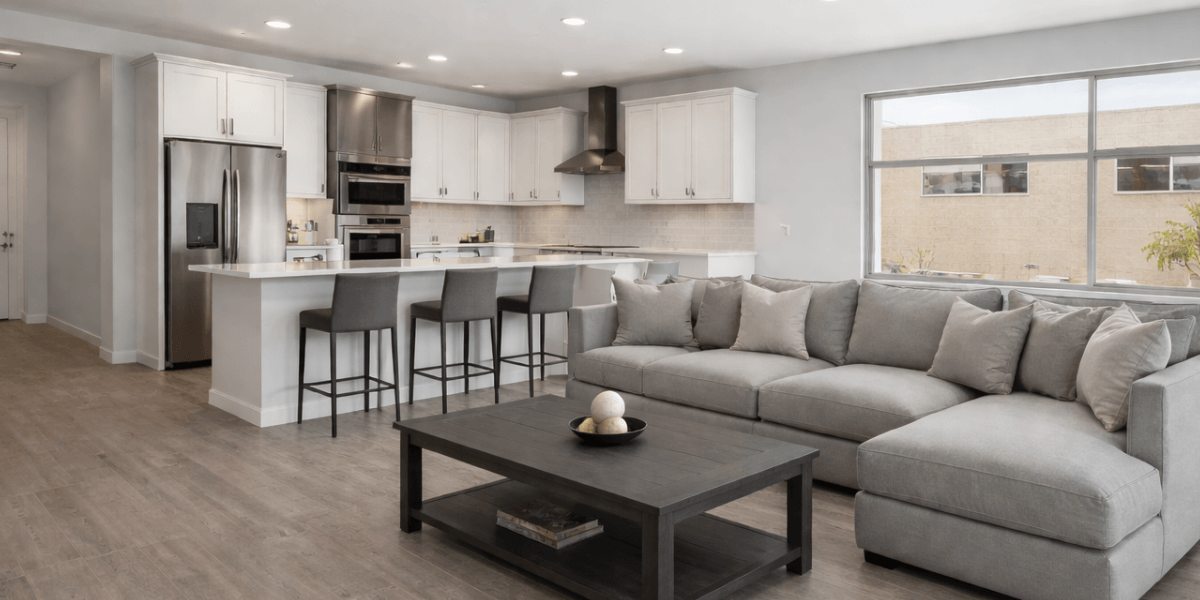 the kitchen and living room at trilogy at wickenburg ranch. gray pallette, an oversized island with bar stools, stainless steel appliances, the kitchen flows into the spacious living room with a large gray sectional couch and coffee table. large windows allow a lot of natural light 
