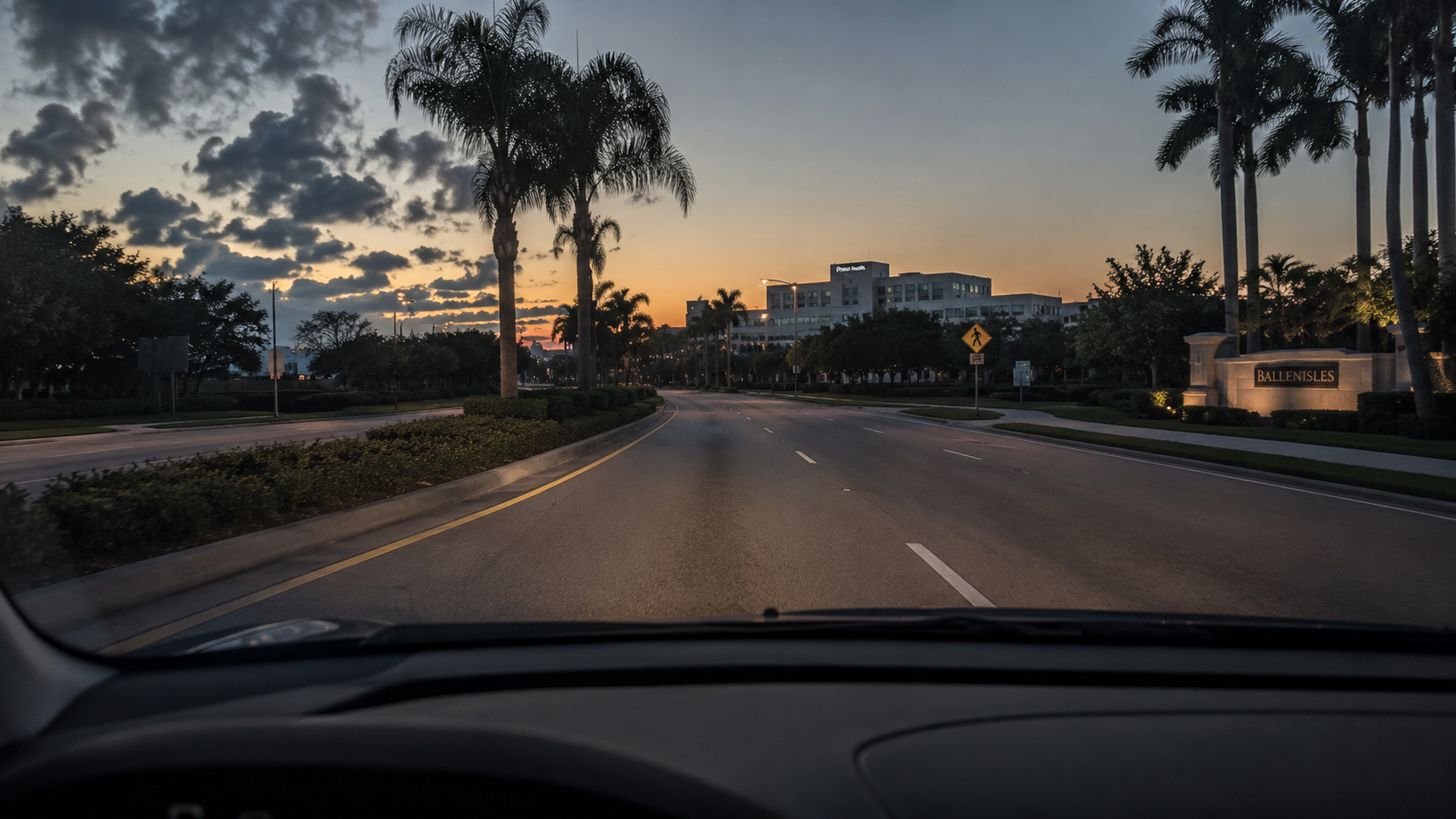 A photorealistic pre-dawn scene captured from the interior of a parked vehicle on a Palm Beach Gardens residential street, with empty lanes stretching toward a faintly visible hospital complex under an orange-gray morning sky. The image supports content addressing commute realities for healthcare workers relocating to Palm Beach County, illustrating the lived experience of shift-hour driving conditions in the Palm Beach Gardens medical corridor. The visual register — quiet, weighted, anticipatory — reflects the broader theme of relocation decisions that reveal themselves through accumulated daily experience rather than in advance.