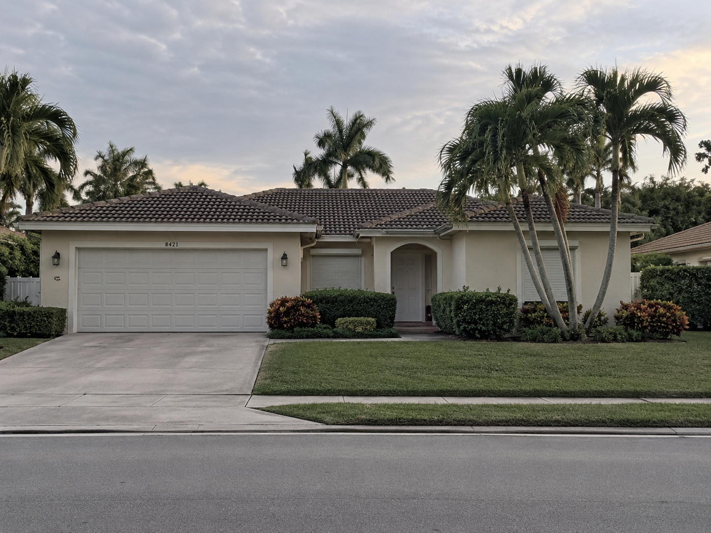A photorealistic exterior of a maintained but unoccupied single-story Florida home in West Palm Beach, FL, captured in flat late-afternoon light with closed shutters and an empty driveway. The scene reflects the quiet legal and emotional weight of an inherited property where heirs have not yet reached agreement on next steps. Intended to support content addressing the question of whether a Florida inherited home can be sold when heirs disagree.