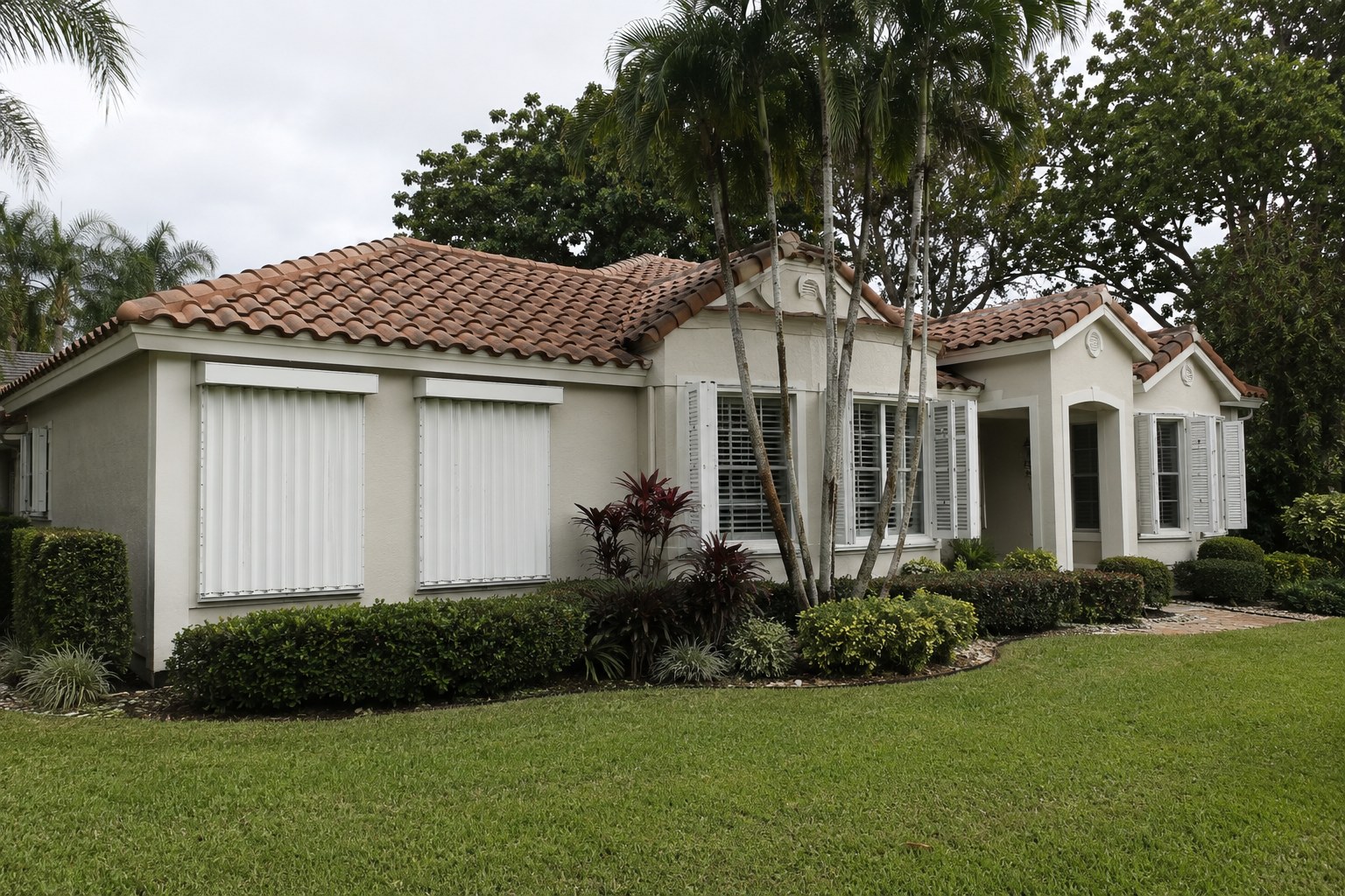 A single-family residence in Palm Beach County, Florida with accordion storm shutters partly deployed and a tile roof in view, photographed in flat overcast light. The scene accompanies an article on homeowners insurance cost for Florida's older homes — what underwriting actually reads in a structure, what an owner can document or improve, and what is locational and therefore fixed.