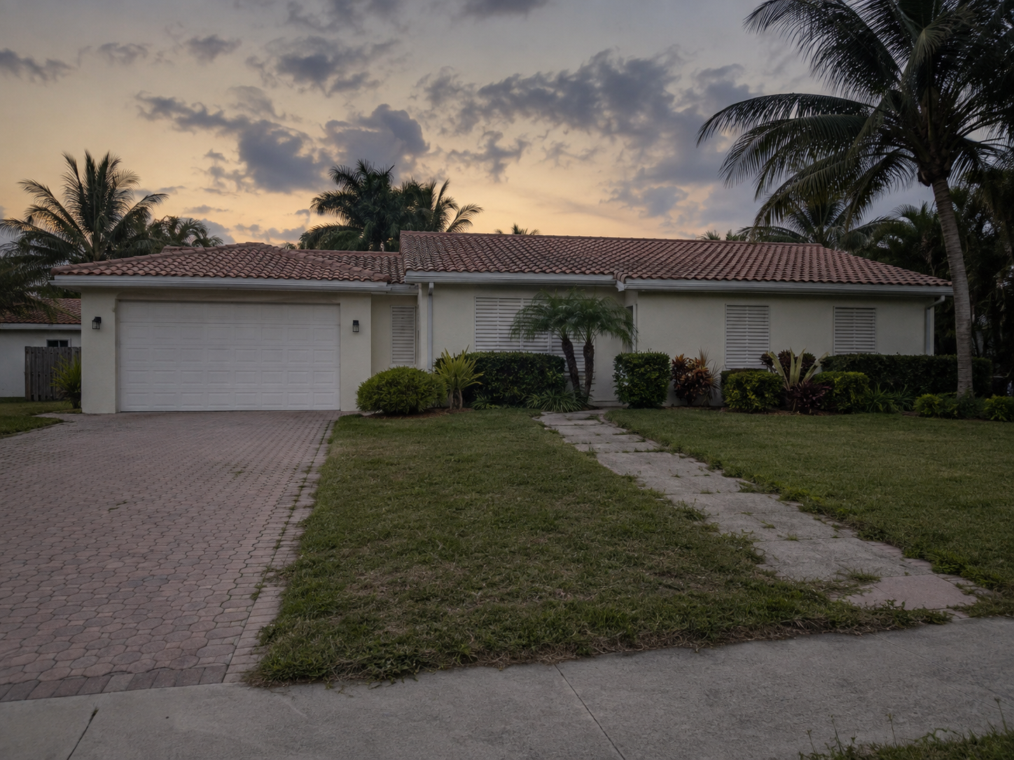 A photorealistic dusk exterior of a single-story Florida residential property in West Palm Beach, shutters closed, interior dark, front path showing light overgrowth — visually representing the suspended state of a multi-heir probate property awaiting resolution. The image contextualizes the coordination challenges that arise when multiple heirs must reach consensus before a Florida probate sale can proceed. Location references West Palm Beach, Palm Beach County, Florida.