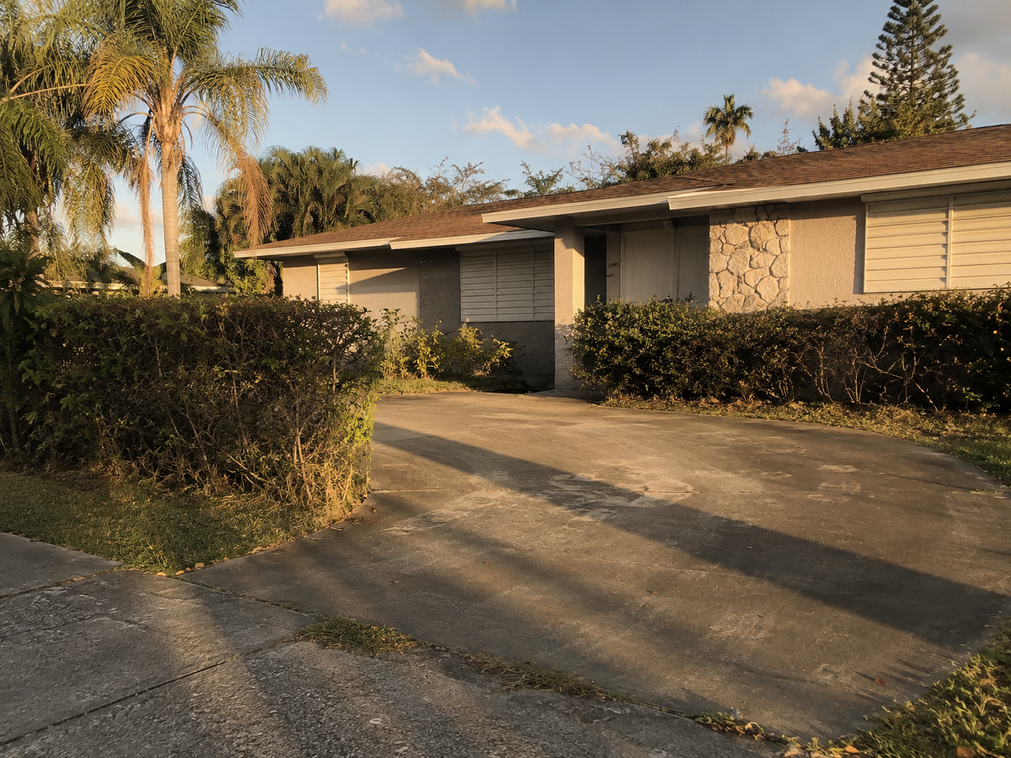 A quiet West Palm Beach residential property photographed in late afternoon light, closed shutters and an overgrown front hedge signaling extended absence. The scene reflects the common condition of Palm Beach County probate properties — maintained minimally, caught between the conclusion of one ownership chapter and the legal steps required to begin the next. The image supports content addressing out-of-state heir misconceptions about Florida probate real estate timelines and authority.