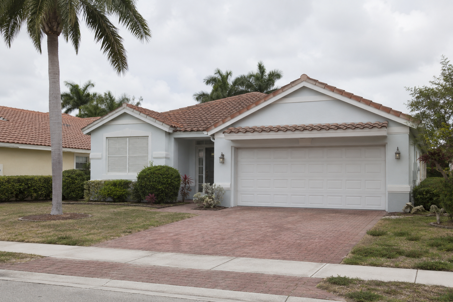 A photorealistic exterior of a single-family home in West Palm Beach, Florida, photographed in flat midday light with shutters half-drawn and an empty driveway. The scene visually represents the quiet accumulation of ownership weight — rising insurance, deferred maintenance decisions, and growing carrying costs — that characterizes the homeowner transition pattern in Palm Beach County's current housing environment. The image is intended to support editorial content on rising housing costs and simplification decisions among South Florida homeowners.