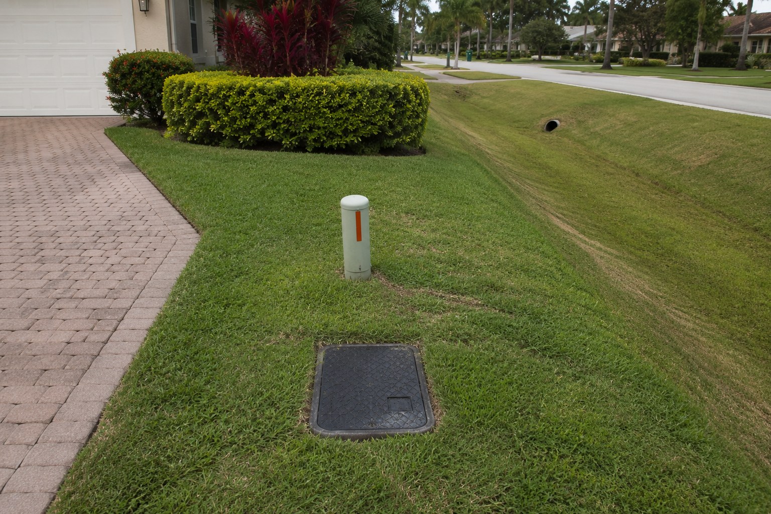Residential utility infrastructure along a lot line in Palm Beach County, Florida — a water meter box, a utility marker post and a drainage swale in flat afternoon light. The scene accompanies an article on utility providers in Palm Beach County by address: how water, sewer, electric, stormwater and waste service are fixed by the parcel rather than selected by the household.