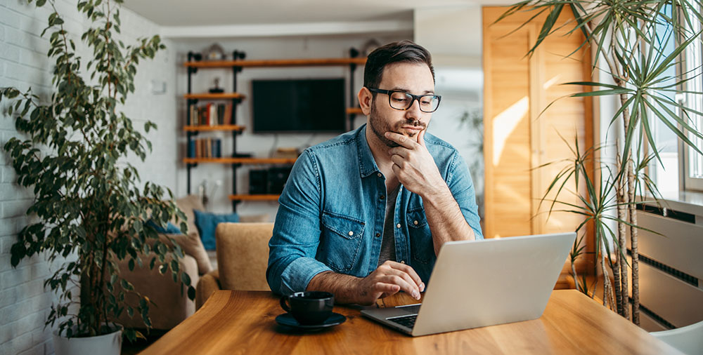 Portrait of a casual adult male thinking while looking at laptop screen, keeping hand on chin.
