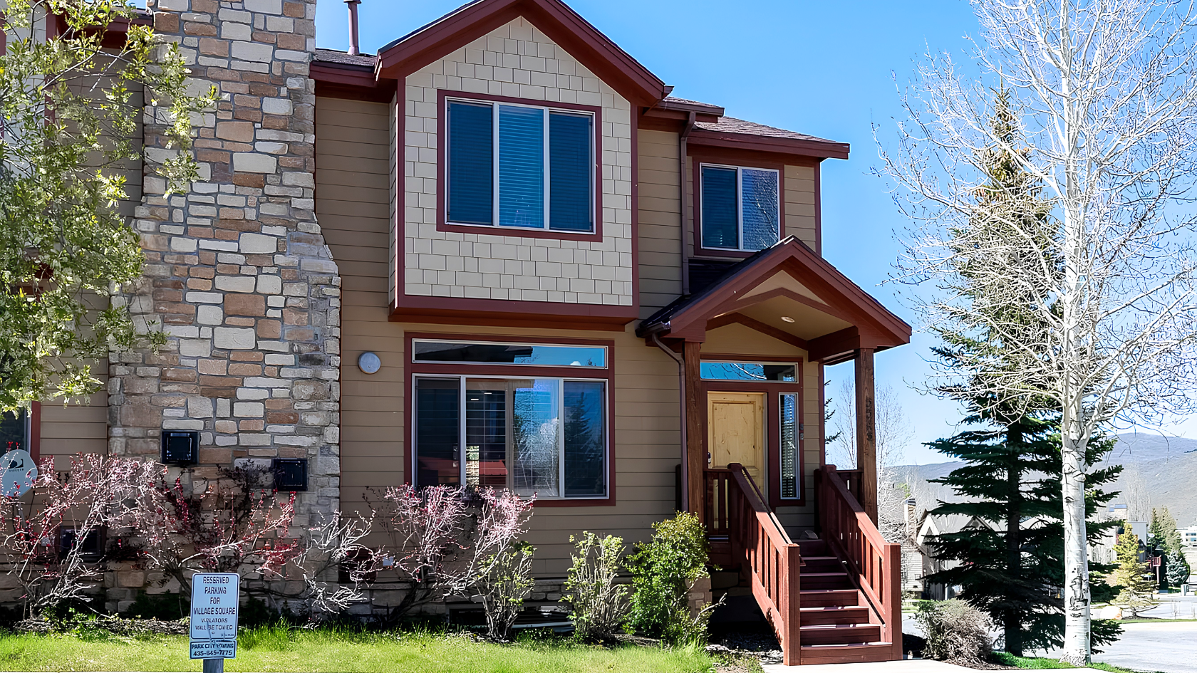 Exterior of 5545 Lillehammer Lane in Park City, Utah &mdash; a two-story mountain condominium with stone accent facade, cedar shingle siding, and red trim, surrounded by mature trees and spring landscaping