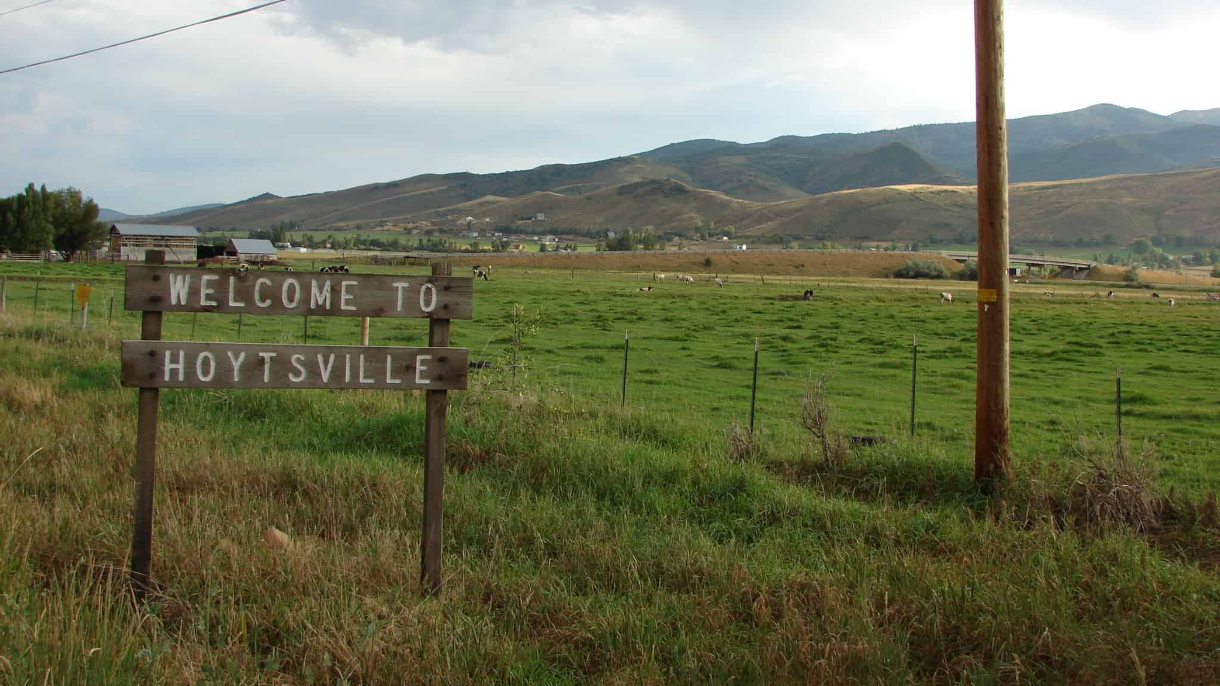Welcome to Hoytsville sign with green pastures and mountain backdrop.