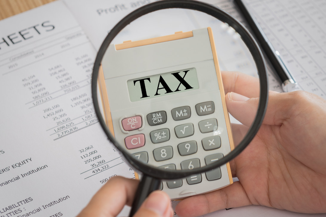 A magnifying glass over a calculator displaying TAX on financial paperwork.